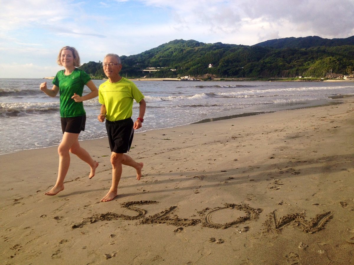 Hiroaki Tanaka in yellow and Magdalena Jackowska in green jogging barefoot on a beach past the word SLOW written in the sand.