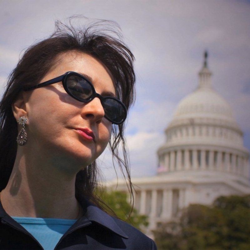 A woman in dark sunglasses and earrings in front of the US Capitol dome.